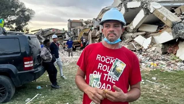 Ronnie Navarro con una franela roja y un casco azul y la pila de escombros atrás.