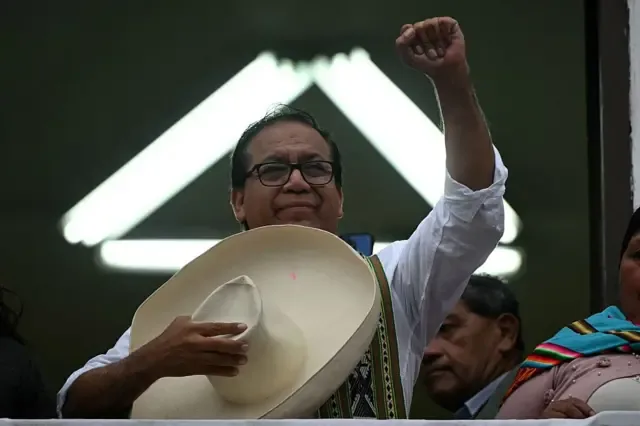 Roberto Sánchez sujeta un gran sombrero con la mano derecha y alza el puño izquierdo. Se ven dos personas junto a él.
